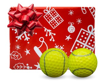 May include: Two bright yellow golf balls with dark grey seam lines positioned in front of a rectangular gift wrapped in red paper featuring a white pattern of presents, snowflakes, holly leaves, and a Santa hat, topped with a red bow.