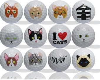 May include: Twelve white golf balls with different cat-themed designs. The designs include a cat with a heart that says "I Love Cats", a cat in a house, a tabby cat, a black cat, a calico cat, a Siamese cat, a gray cat, a brown cat, and a white cat with green eyes.