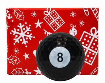 May include: A black golf ball featuring a white circular center with the number 8 printed in black, positioned in front of red wrapping paper decorated with white holiday patterns including snowflakes, gift boxes, and ornaments.