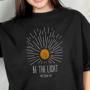 Puede incluir: Una camiseta negra con un gráfico blanco de un sol con rayos y el texto "Be the Light Matthew 5:14".