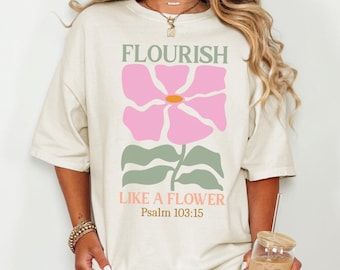 May include: Oversized cream t-shirt featuring a pink flower graphic and the text "FLOURISH LIKE A FLOWER Psalm 103:15". The shirt is paired with denim shorts. The overall style is casual and relaxed.