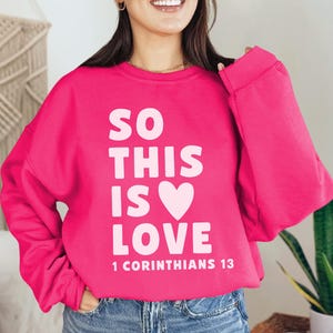 May include: A bright pink sweatshirt with the text "SO THIS IS LOVE" in white, with a white heart symbol. The text "1 Corinthians 13" is printed below. The sweatshirt is a crew neck style.
