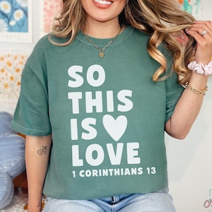 May include: Sage green t-shirt with the text "SO THIS IS LOVE" and "1 CORINTHIANS 13" in white. The shirt features a white heart graphic. The model is wearing blue jeans and a gold necklace.