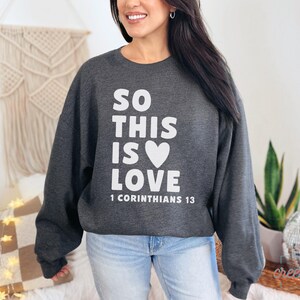 May include: Dark gray sweatshirt with the text "SO THIS IS LOVE" in white, a white heart, and the Bible verse "1 Corinthians 13". The sweatshirt is a crew neck style.