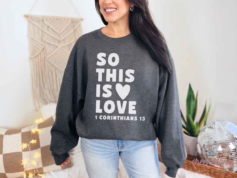May include: Dark gray sweatshirt with the text "SO THIS IS LOVE" in white, a white heart, and the Bible verse "1 Corinthians 13". The sweatshirt is a crew neck style.