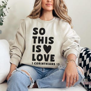 May include: Beige sweatshirt with the text "SO THIS IS LOVE" in black, with a black heart and the Bible verse "1 Corinthians 13". The sweatshirt is paired with ripped blue jeans.