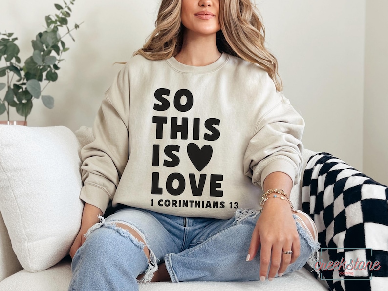 May include: Beige sweatshirt with the text "SO THIS IS LOVE" in black, with a black heart and the Bible verse "1 Corinthians 13". The sweatshirt is paired with ripped blue jeans.