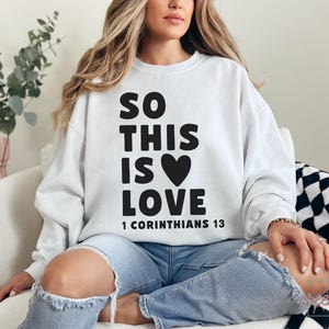 May include: White sweatshirt with the text "SO THIS IS LOVE" and a black heart graphic. The text "1 CORINTHIANS 13" is printed below. The sweatshirt is worn with blue jeans.