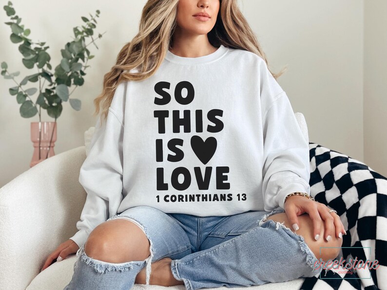 May include: White sweatshirt with the text "SO THIS IS LOVE" and a black heart graphic. The text "1 CORINTHIANS 13" is printed below. The sweatshirt is worn with blue jeans.