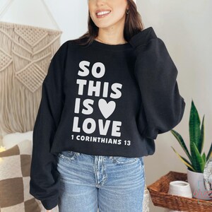 May include: Black sweatshirt with the text "SO THIS IS LOVE" and a white heart graphic. The text "1 Corinthians 13" is below the main text. The sweatshirt is paired with blue jeans.