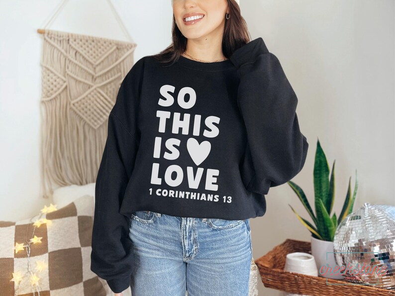 May include: Black sweatshirt with the text "SO THIS IS LOVE" and a white heart graphic. The text "1 Corinthians 13" is below the main text. The sweatshirt is paired with blue jeans.