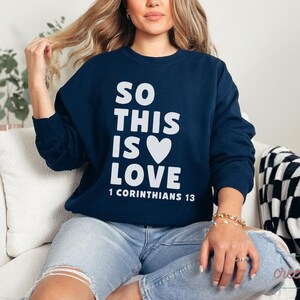 May include: Navy blue sweatshirt with the text "SO THIS IS LOVE" in white, along with a white heart graphic. The text "1 CORINTHIANS 13" is below. The sweatshirt is long-sleeved and made of a soft material.