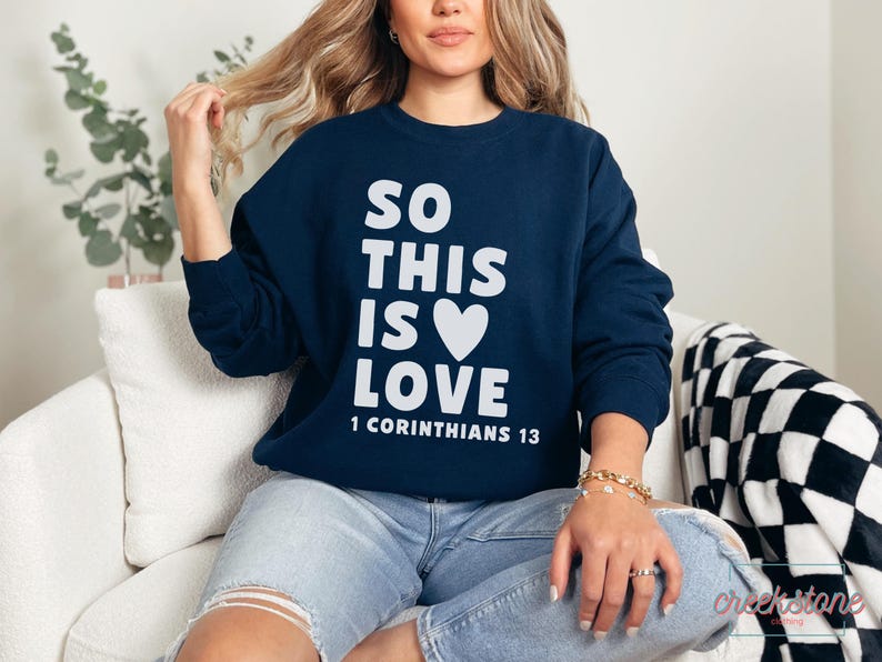 May include: Navy blue sweatshirt with the text "SO THIS IS LOVE" in white, along with a white heart graphic. The text "1 CORINTHIANS 13" is below. The sweatshirt is long-sleeved and made of a soft material.