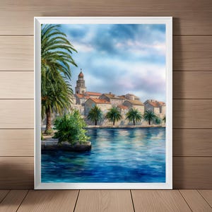 May include: Watercolor painting of a coastal town with a church tower, palm trees, and buildings with red tile roofs. The artwork is framed in white and depicts a serene scene with blue water and a cloudy sky.