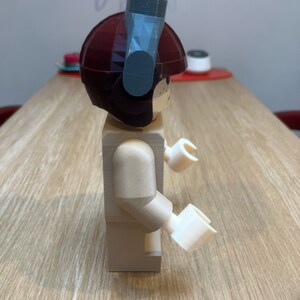 Puede incluir: Una minifigura Lego beige con un casco marr&oacute;n y un accesorio gris en la parte superior. La minifigura tiene brazos y manos blancos.