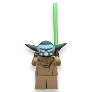 May include: A green and brown Lego minifigure of Yoda from Star Wars holding a green lightsaber. The minifigure is facing away from the camera.