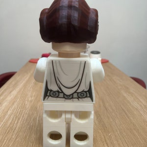 Puede incluir: Una figura de Lego blanca de la princesa Leia de Star Wars. La figura lleva su caracter&iacute;stico vestido blanco y tiene una peluca marr&oacute;n. La figura est&aacute; de pie sobre una superficie de madera.