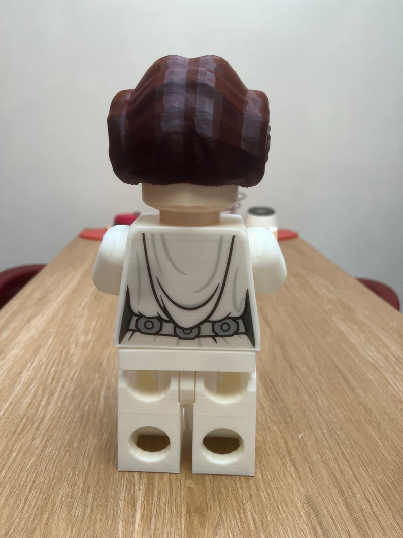 Puede incluir: Una figura de Lego blanca de la princesa Leia de Star Wars. La figura lleva su caracter&iacute;stico vestido blanco y tiene una peluca marr&oacute;n. La figura est&aacute; de pie sobre una superficie de madera.