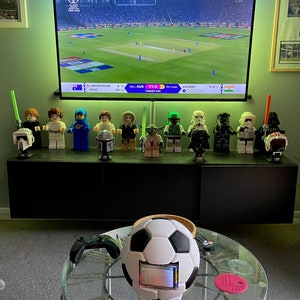 May include: A collection of Star Wars action figures, including Darth Vader, Yoda, and a Stormtrooper, are displayed on a shelf in front of a television screen showing a cricket match. The score is 71-3, with Australia batting. A football with a screen embedded in it sits on a glass table with a pink coaster and a wireless charging pad.