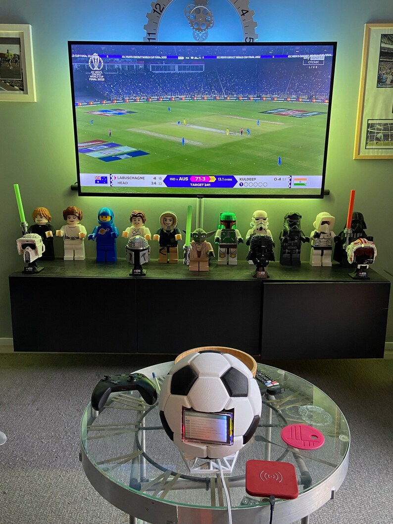 May include: A collection of Star Wars action figures, including Darth Vader, Yoda, and a Stormtrooper, are displayed on a shelf in front of a television screen showing a cricket match. The score is 71-3, with Australia batting. A football with a screen embedded in it sits on a glass table with a pink coaster and a wireless charging pad.