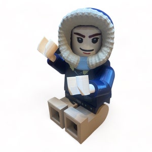 May include: A 3D-printed figurine of a person in a blue parka with a white fur-lined hood. The figure is holding two white objects and is seated on a brown sled-like base. The figure has a smiling expression.