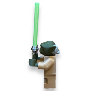 May include: A green and brown Lego minifigure of Yoda holding a green lightsaber. The minifigure is standing on a white background.