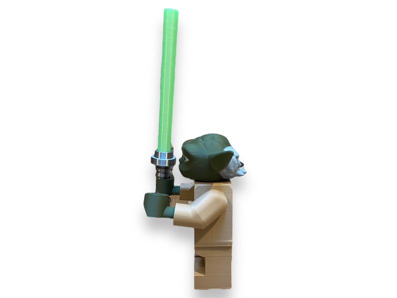 May include: A green and brown Lego minifigure of Yoda holding a green lightsaber. The minifigure is standing on a white background.