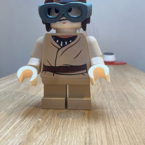 Puede incluir: Una minifigura Lego de color marr&oacute;n y beige que lleva un casco marr&oacute;n con una franja azul y gafas verdes. La minifigura lleva una camisa beige con una franja negra y un cintur&oacute;n marr&oacute;n.
