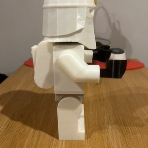 May include: A white 3D printed Lego-style figure of a Stormtrooper from Star Wars. The figure is wearing a helmet and has a backpack. The figure is holding a black weapon in its right hand.