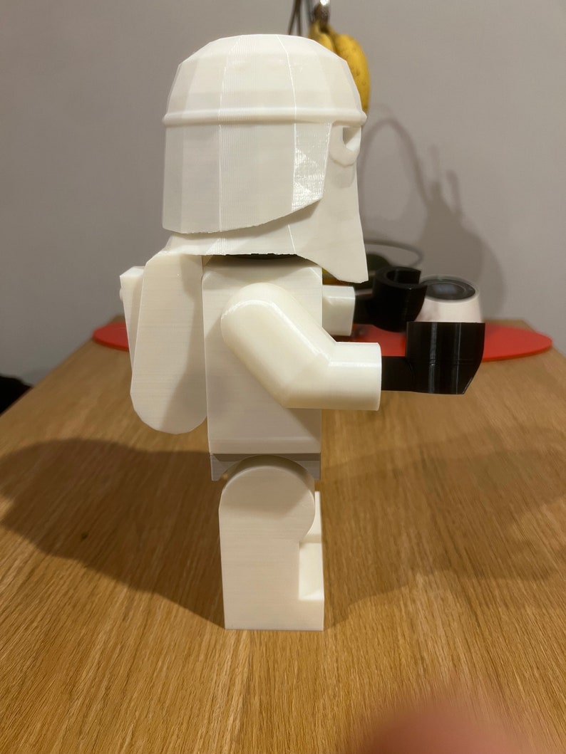 May include: A white 3D printed Lego-style figure of a Stormtrooper from Star Wars. The figure is wearing a helmet and has a backpack. The figure is holding a black weapon in its right hand.