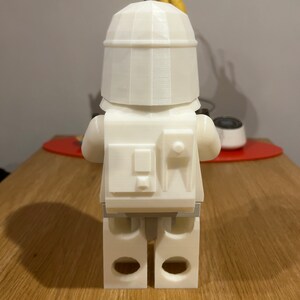 May include: White plastic figurine of a Lego character wearing a helmet and a backpack. The figurine is standing on a wooden surface.