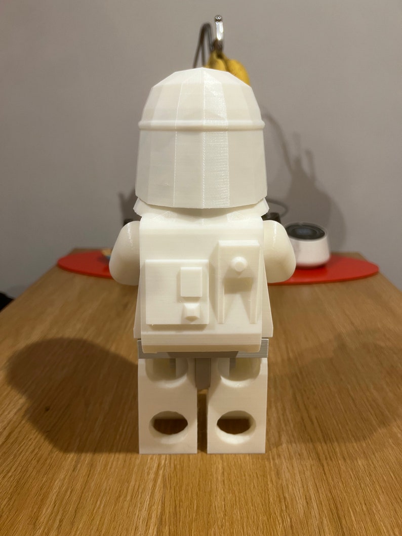 May include: White plastic figurine of a Lego character wearing a helmet and a backpack. The figurine is standing on a wooden surface.