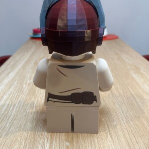 Puede incluir: Una minifigura Lego beige con un cintur&oacute;n marr&oacute;n y un casco. El casco est&aacute; hecho de pl&aacute;stico azul y rojo y tiene un dise&ntilde;o geom&eacute;trico.
