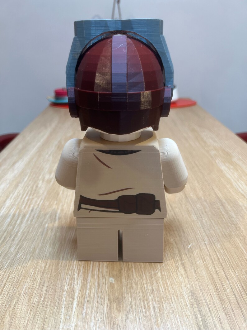 Puede incluir: Una minifigura Lego beige con un cintur&oacute;n marr&oacute;n y un casco. El casco est&aacute; hecho de pl&aacute;stico azul y rojo y tiene un dise&ntilde;o geom&eacute;trico.