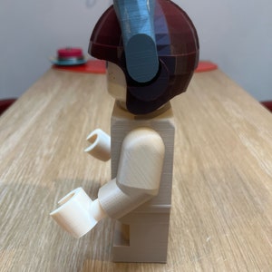 Puede incluir: Una figura de Lego beige con un casco marr&oacute;n y un accesorio azul y gris en la parte superior. La figura est&aacute; de pie sobre una superficie de madera.