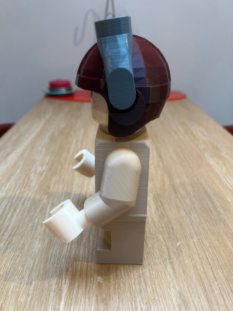 Puede incluir: Una figura de Lego beige con un casco marr&oacute;n y un accesorio azul y gris en la parte superior. La figura est&aacute; de pie sobre una superficie de madera.
