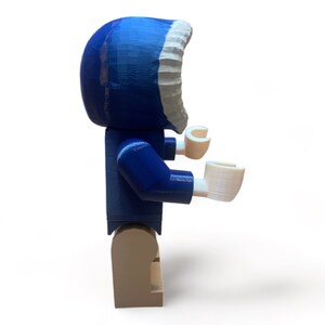 May include: A blue and white Lego-style figure with a hooded parka. The figure has white hands and tan legs. The parka hood is blue with a white trim. The figure is standing on a white surface.