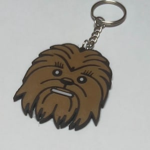 May include: A brown rubber keyring featuring the stylised face of a Wookiee character with dark brown outlines, white eyes, and a white mouth. The keyring is attached to a standard silver-coloured metal split ring and a short chain link.