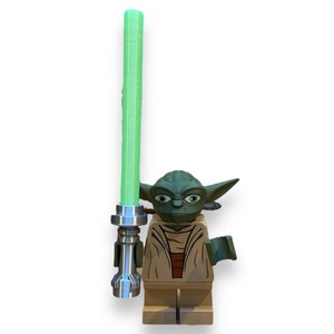 May include: A green and brown Lego minifigure of Yoda holding a green lightsaber. The lightsaber has a silver handle and a green blade.