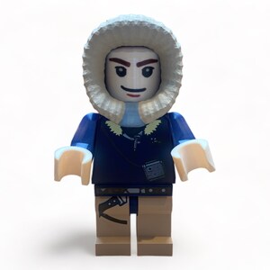 May include: A Lego minifigure of a person wearing a blue jacket with a fur-lined hood. The figure has tan legs, light tan hands, and a small bag. The figure is standing against a white background.