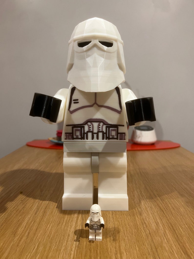 May include: A large white LEGO Star Wars Snowtrooper figure stands next to a smaller, regular-sized LEGO Snowtrooper figure. Both figures are wearing white helmets and armour.