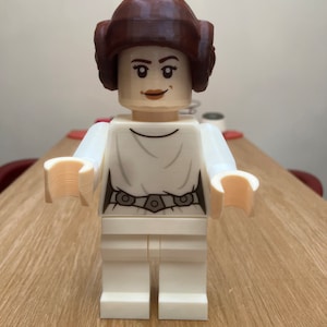 Puede incluir: Una figura de Lego blanca de la princesa Leia de Star Wars. Tiene una peluca marr&oacute;n y un vestido blanco con un cintur&oacute;n negro.