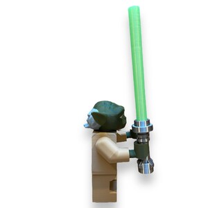 May include: A green and brown Yoda Lego minifigure holding a green lightsaber. The lightsaber has a silver handle and a green blade.