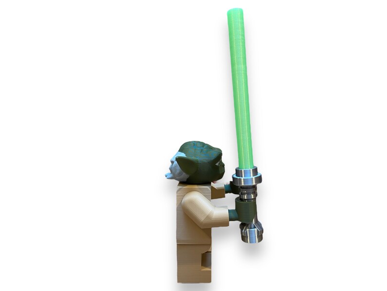 May include: A green and brown Yoda Lego minifigure holding a green lightsaber. The lightsaber has a silver handle and a green blade.