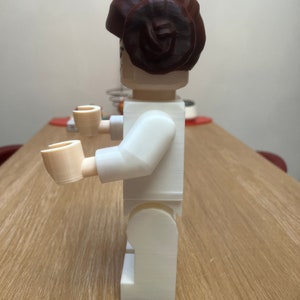 Puede incluir: Una figura de Lego de pl&aacute;stico blanco de una mujer con un mo&ntilde;o de pelo marr&oacute;n. La figura est&aacute; de pie sobre una superficie de madera.