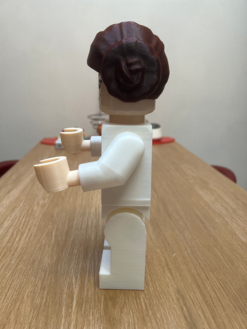 Puede incluir: Una figura de Lego de pl&aacute;stico blanco de una mujer con un mo&ntilde;o de pelo marr&oacute;n. La figura est&aacute; de pie sobre una superficie de madera.