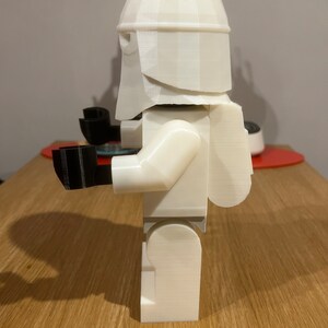 May include: White 3D printed Lego-style figure of a Star Wars Stormtrooper. The figure is standing on a wooden surface.