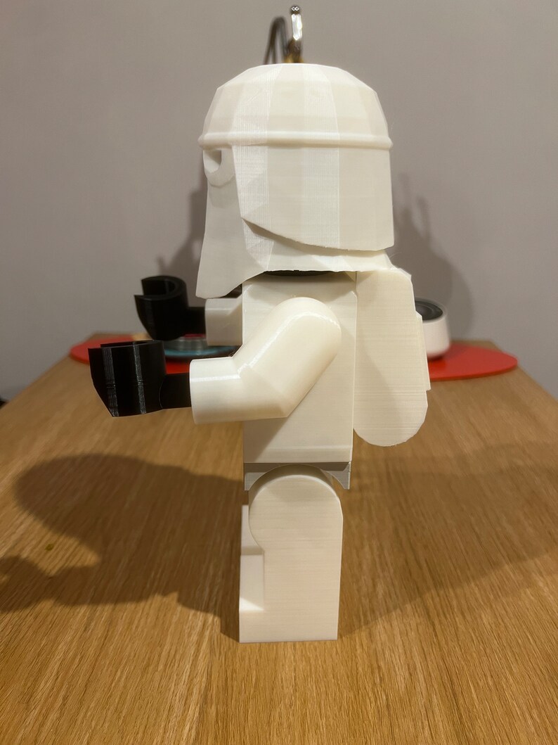 May include: White 3D printed Lego-style figure of a Star Wars Stormtrooper. The figure is standing on a wooden surface.