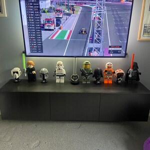 May include: A black entertainment center with a large television displaying a Formula 1 race. The TV is mounted above a row of Lego Star Wars character figures. A soccer ball and remote control are in the foreground.