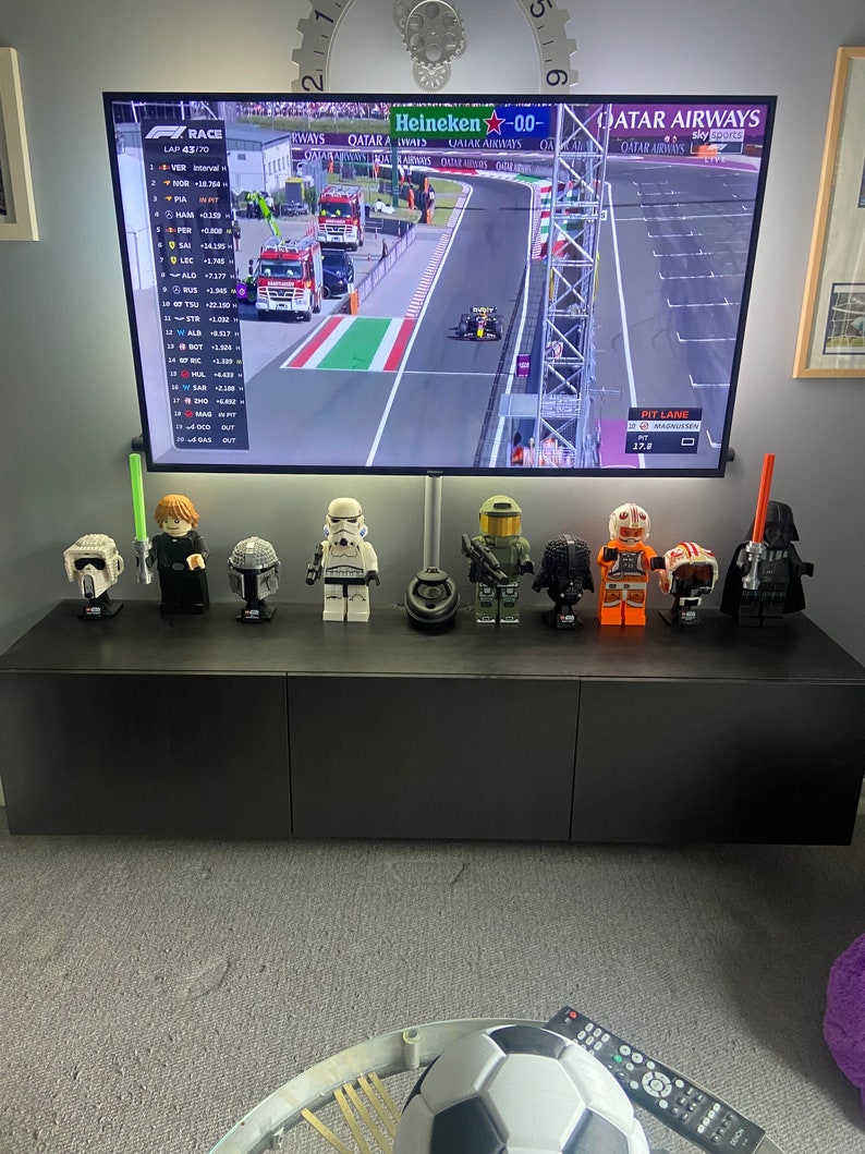 May include: A black entertainment center with a large television displaying a Formula 1 race. The TV is mounted above a row of Lego Star Wars character figures. A soccer ball and remote control are in the foreground.